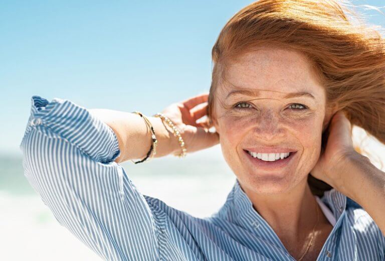 Portrait of beautiful mature woman with wind fluttering hair. Closeup face of healthy young woman with freckles relaxing at beach. Cheerful lady with red hair and blue blouse standing at seaside enjoying breeze looking at camera. 